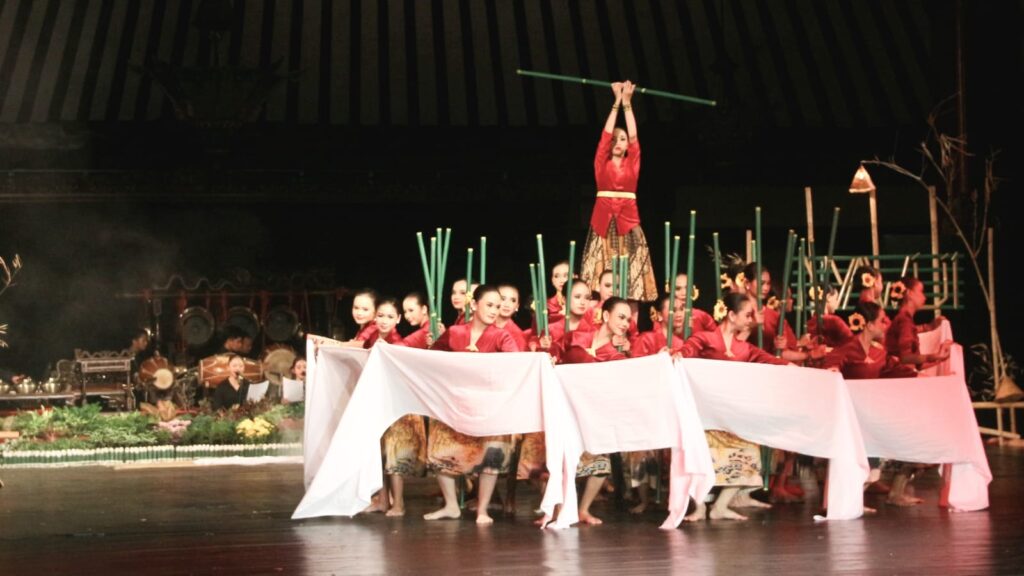 Dancers in red outfits perform a bamboo stick routine on stage with one performer elevated and holding a green pole above the crowd